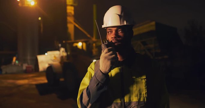 African foreman with walkie talkie supervising night shift work of asphalt manufacturing