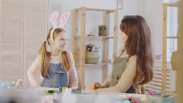 Medium shot of cheerful young woman and cute preteen girl smiling and putting bunny ears on each other, then taking unpainted Easter bunny figurine and deciding on how to decorate it
