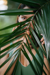 young caucasian lady in green swimsuit covering face with exotic green plant, only eyes are visible behind plant. summer, resort and holiday concept. green texture