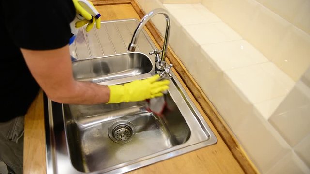 Caucasian man cleaning iron sink with spray, spoonge and gloves