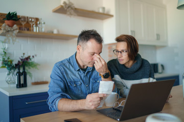 Young stressed Caucasian couple facing financials troubles, sitting at kitchen table with bills, checks and laptop computer and reading document from bank, looking frustrated and unhappy