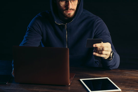 cropped caucasian hacker man with beard holding card in hand, wearing pullover, sit in the hood and studying bank card, try to find information and hack, male commiting a crime using laptop and tablet