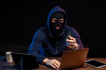 aggressive anonymous hacker man in mask sit with laptop isolated in dark space, man uses computers...
