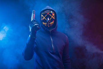 confident dangerous killer male in pullover stand in the hood with gun, anonymous masked hunter man isolated over smoky space with neon lights. crime, kill, hunt, danger concept - obrazy, fototapety, plakaty