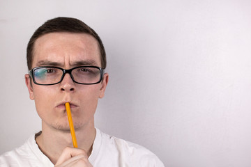 Guy wearing a white T-shirt and transparent glasses holds a yellow pencil by his lips, on gray background.