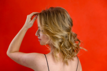 Fototapeta premium The concept of cosmetics, fashion and style. Glamorous beauty rear view portrait of a pretty model with blond hair with great makeup and a beautiful hairstyle on a red background in the studio.