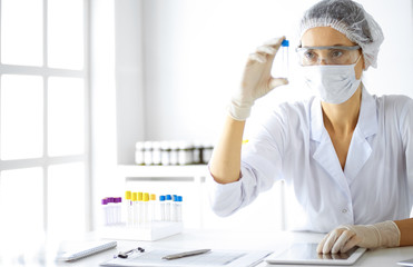 Professional female scientist in protective eyeglasses researching tube with reagents in...