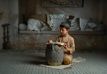 Young muslim boy in prayer cap and arabic clothes with rosary beads reading holy quran book praying...