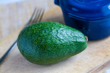 A ripe avocado on a wooden board, a blue baking pot, and a fork