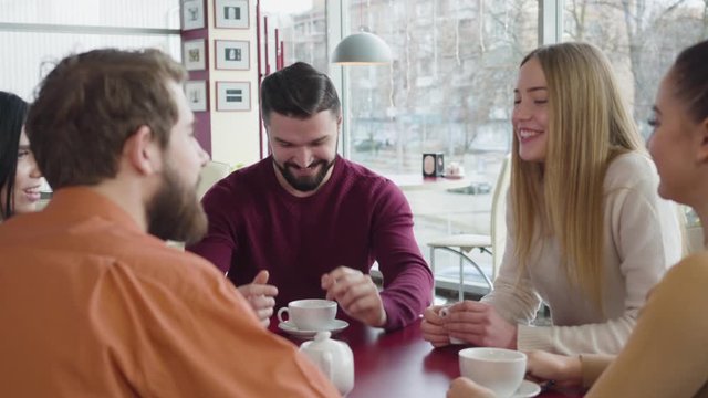 Portrait of handsome bearded Caucasian man talking with group of friends in cafe and smiling. Positive young man telling stories and having fun with fellows. Lifestyle, joy, leisure.
