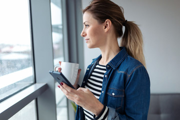 Pretty young woman using her mobile phone while drinking coffee near to the window in the living room at home.