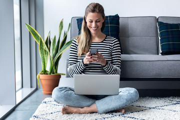 Pretty young woman using her mobile phone while working with laptop sitting on the floor at home.