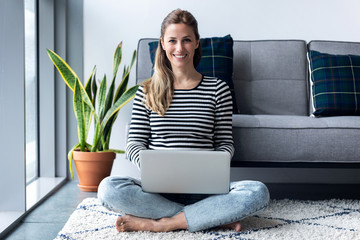 Pretty young woman looking at camera while using her laptop at home.