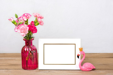 Small colored pink carnations in a vase and white photo frame, a figure of flamingo on wooden background and grey wall Hello spring, seasonal concept Mock up Place for text
