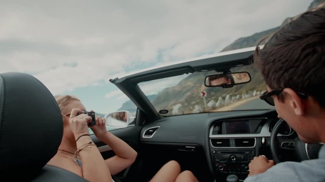 Young couple on a road trip. Man driving car with woman taking pictures with a digital camera.