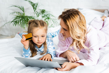 Fototapeta na wymiar mother looking at laptop and daughter holding credit card in bedroom