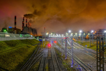 Fototapeta na wymiar A network of intertwined railroad tracks illuminated by bright illumination at night and pipes of industrial enterprises that spew puffs of smoke into the atmosphere. Industrial landscape showing terr