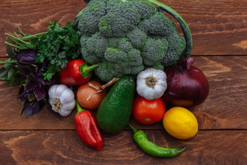 Vegetables, broccoli, chili pepper, bell pepper, onion, garlic, avocado, tomato, lemon and a bunch of oregano and parsley lie on a brown wooden kitchen table, top view, rustic, flat lay