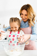 Obraz na płótnie Canvas Selective focus of attractive mom and cute daughter smiling and mixing ingredients for tasty cupcakes together