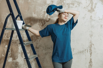 A young woman in work clothes wearily wipes sweat from her forehead after preparing the plastered wall for painting.