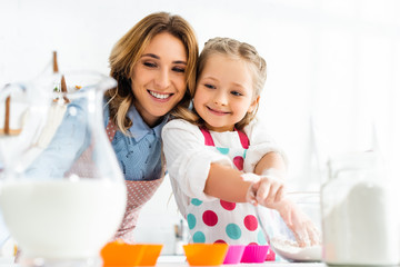 Selective focus of beautiful mother and daughter preparing cupcakes with ingredients including milk and flour in kitchen