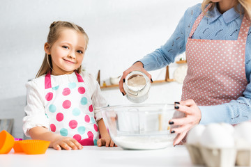 cropped view of mother adding flour to bowl and smiling daughter looking at camera