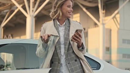Cheerful blonde business woman in coat using smartphone and looking away while standing near the car on airport parking outdoors - Powered by Adobe