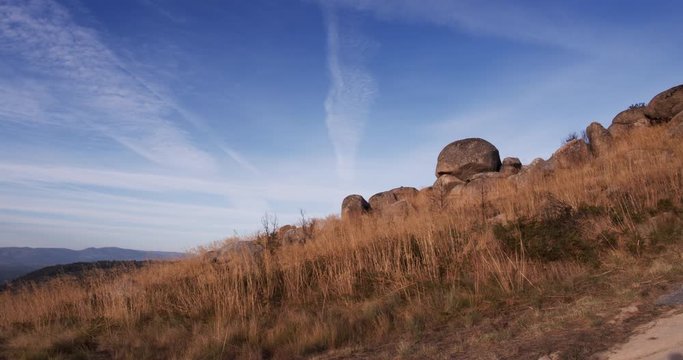 Landscape field of dry grass on a mountain hill timelapse. Clouds moving fast and hay waving in the wind near a pre historical huge rock.