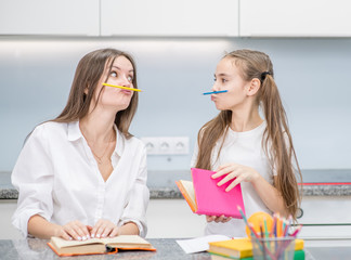 Young mother and her daughter are sitting at the table and having fun and foolish while doing homework together