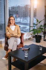 caucasian happy woman psychologist sit on leather chair in her own office and laugh. cheerful lady look side, enjoy working as psychology, love to help people. big panoramic window in the background