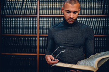Smart male bookworm 30s making research in public library standing near publicity area, concentrated young professor reading articles in encyclopedia choosing literature for university project