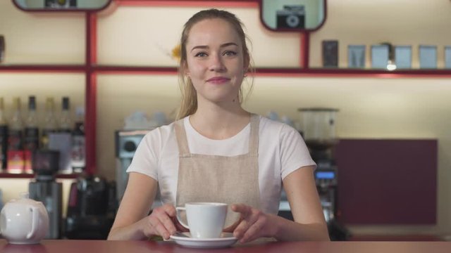 Portrait of cheerful young Caucasian woman putting coffee cup on the table, looking at camera and smiling. Happy waitress working in cafe. Lifestyle, joy,. occupation, satisfaction.