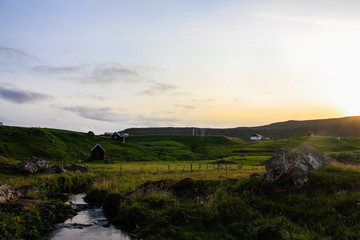 Fototapeta premium Green hilly meadow and small houses on it. Evening sunset. Sunlight.