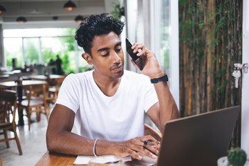 Wistful male typing on laptop and communicating on mobile in eatery