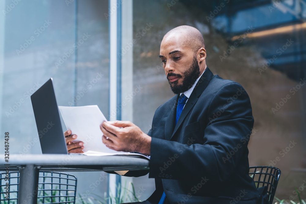 Wall mural Bald ethnic worker reading report in front of laptop