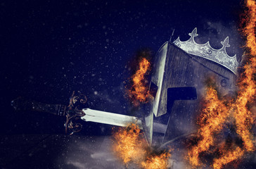 photo of crown over king knight helmet armor and sword in front of fire flames and dark background. Medieval period concept