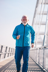 A senior sportsperson running on a bridge walkway, cheerful and smiling. He is wearing blue sports clothes.