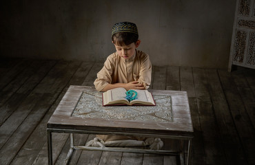 Fototapeta na wymiar Young muslim boy in prayer cap and arabic clothes with rosary beads reading holy quran book praying to Allah, ramadan kareem concept kid spiritual peaceful moment inside eastern traditional interior