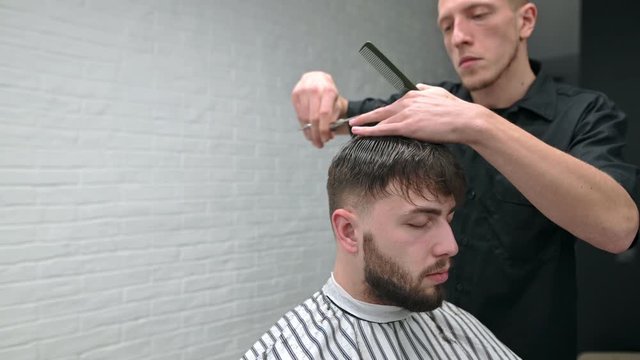 Barber in a black shirt cuts hair with scissors to bearded client.Bearded man sits in a barber shop with his eyes closed and cuts his hair at a male hairdresser. Barber creates a stylish men's haircut