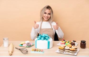 Teenager pastry chef with a big cake in a table giving a thumbs up gesture