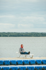 A girl in dark blue jeans and a gray warm jacket is sitting on a sunbed on a pontoon, against the background of a vast lake and forest. She's meditating