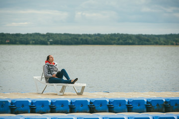 A girl in dark blue jeans and a gray warm jacket is sitting on a sunbed on a pontoon, against the...