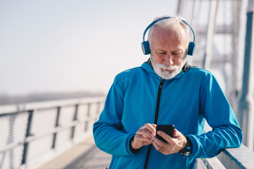 An elderly man on a bridge walkway wearing headphones and holding his phone.