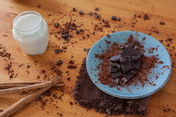 Ounces of healthy chocolate with pure cocoa on a blue plate, with a wooden spoon with cocoa, a jar of oat milk and cinnamon sticks with a rustic brown cloth and a wooden cloth background