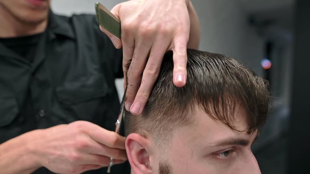 Close up shot, hands of hairdresser with scissors and comb create stylish hairstyle for bearded man. Process of the barber. Male hairdresser cuts the client, uses scissors.