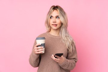 Teenager blonde girl wearing a sweater over isolated pink background holding coffee to take away and a mobile while thinking something
