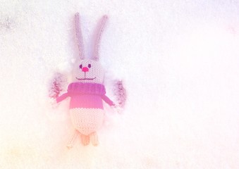 A gray knitted toy hare lies on the snow, enjoys life and waves his paws.