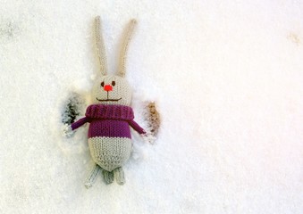 A gray knitted toy hare lies on the snow, enjoys life and waves his paws.
