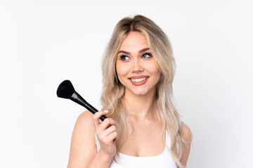 Teenager blonde girl over isolated white background holding makeup brush and looking up