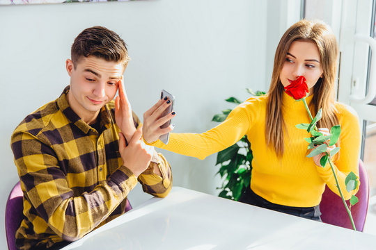 Annoyed boy waiting, looking disapprovingly, while his dirlfriend is making selfie with red rose he brought her.